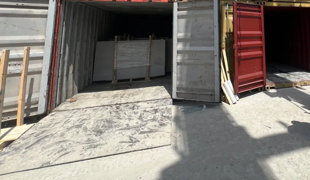 Open shipping containers in a loading yard, one holding an upright stone package.