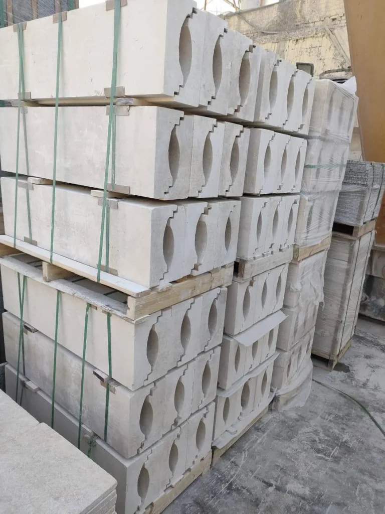 Stacks of shaped pale-stone components with repeating oval recesses, secured on wooden pallets.