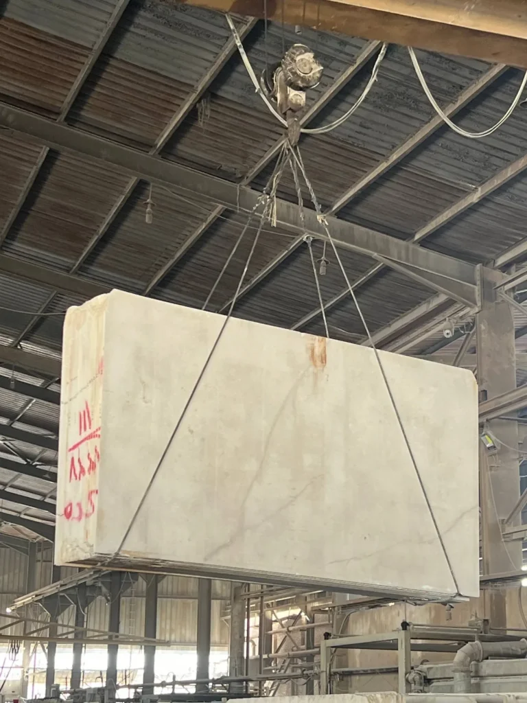 Large pale stone block suspended by lifting cables inside a metal-roofed industrial hall.