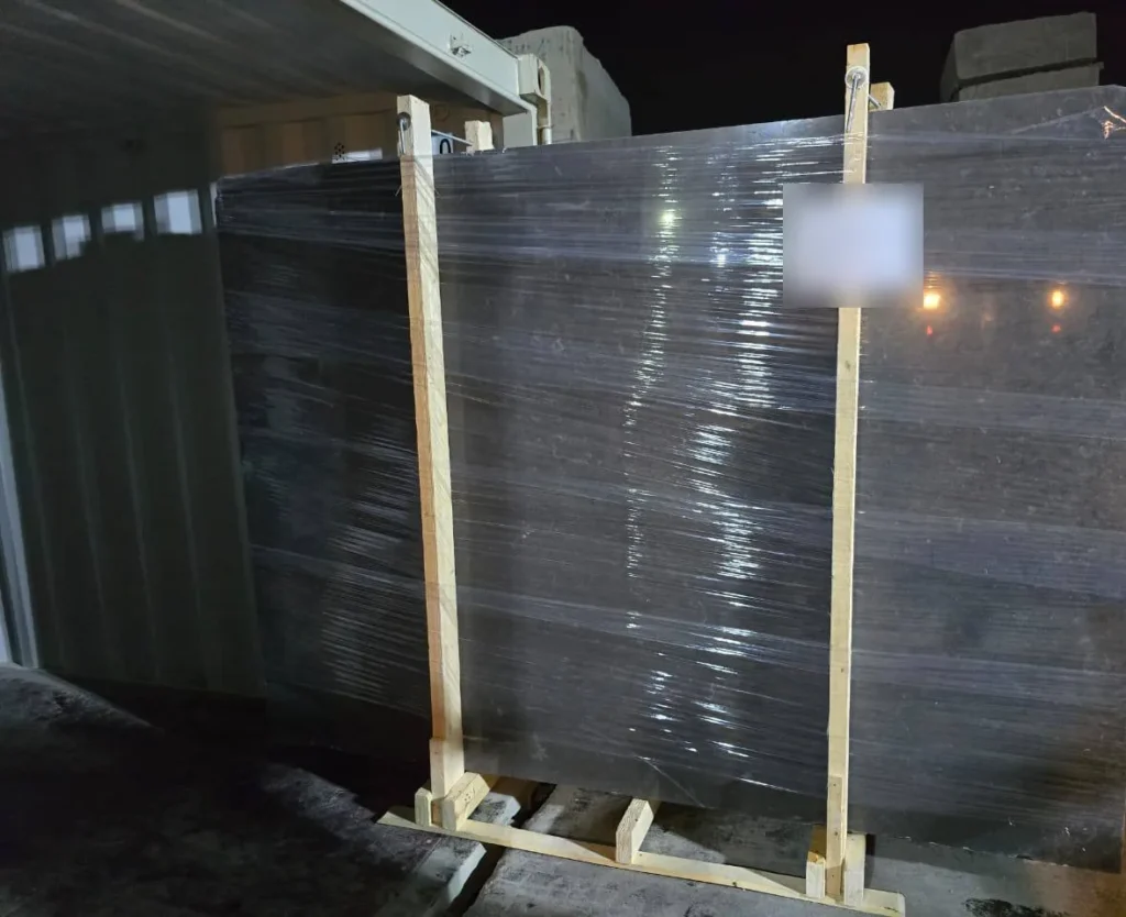 Large dark stone slab bundle wrapped in clear film and held between two timber uprights beside a container.