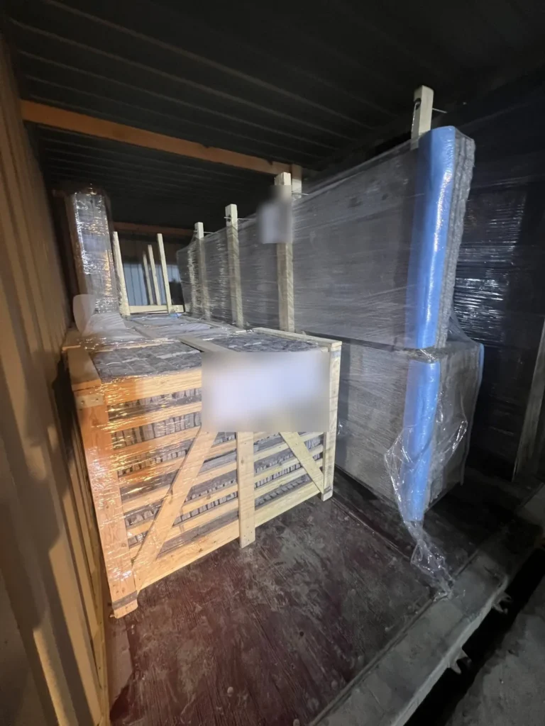 Inside a container: a timber crate of stone pieces beside upright wrapped slab bundles with blue edge protection.